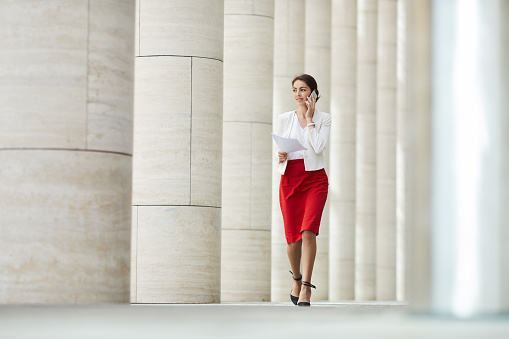 Full length portrait of beautiful woman speaking by phone while walking towards camera along row of pillars, copy space