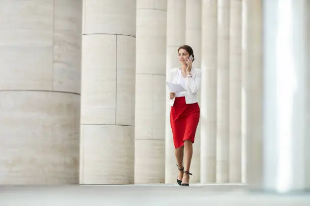 Full length portrait of beautiful woman speaking by phone while walking towards camera along row of pillars, copy space