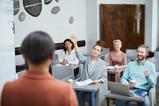 Wide angle portrait of contemporary young woman raising hand in class with group of student enjoying lecture, teacher point of view
