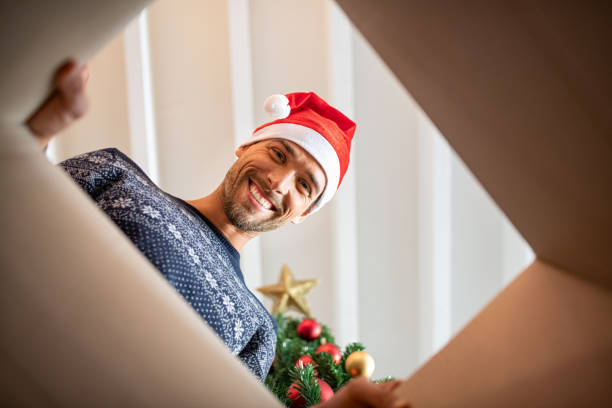 Young man unpacking christmas present Happy man wearing christmas hat unpacking xmas gift. Excited guy smiling while opening present and looking inside. Portrait of handsome guy opening carboard box under the christmas tree while looking at camera. unwrapping stock pictures, royalty-free photos & images