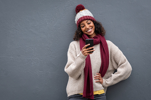 Young african woman using mobile phone standing against grey wall. Smiling beautiful girl wearing warm clothes sending message from cellphone. Black woman wearing sweater and knitted hat while surfing the net with smartphone isolated on gray background with copy space.