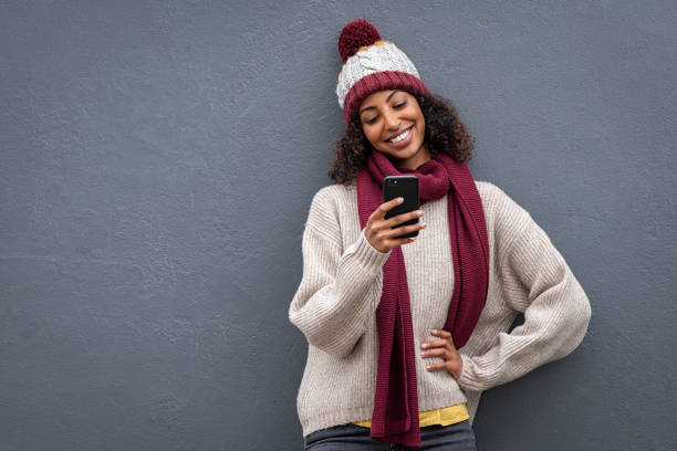 mulher preta nova na roupa do inverno usando o smartphone - scarf women hat warm clothing - fotografias e filmes do acervo