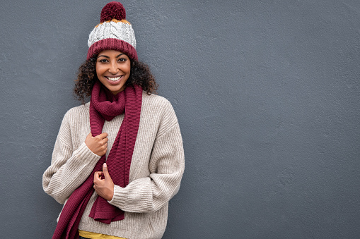 Beautiful young black woman in winter clothing standing and leaning against grey wall. Portrait of attractive african woman wearing woolen cap and scarf and looking at camera. Smiling girl feeling cold on gray background.
