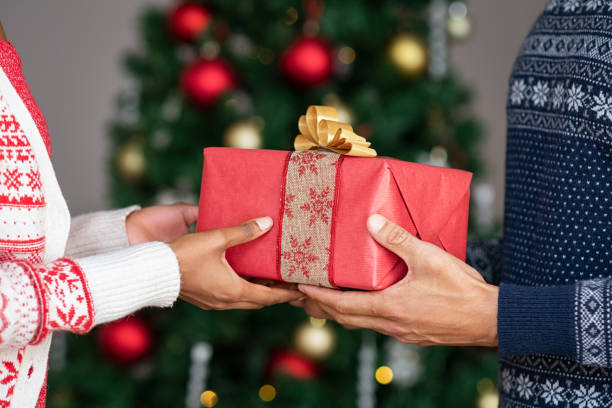 Hands giving christmas gifts Couple wearing christmas theme sweater exchanging gifts in front of decorated christmas tree. Closeup of african woman hand receive xmas present from her boyfriend. Young couple holding christmas gift. christmas human hand christmas ornament decoration stock pictures, royalty-free photos & images