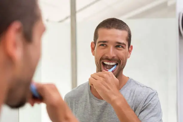 Smiling young man with toothbrush cleaning teeth and looking mirror in the bathroom. Handsome young man brushing his teeth in morning in bathroom. Happy guy in pajamas brushing teeth at night before going to sleep.