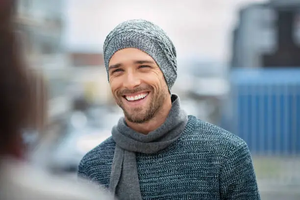 Young man wearing winter hat and scarf laughing while talking to woman. Handsome guy standing on a sidewalk wearing jumper and cashmere scarf. Portrait of cheerful man in winter clothes enjoying outdoor with girlfriend.