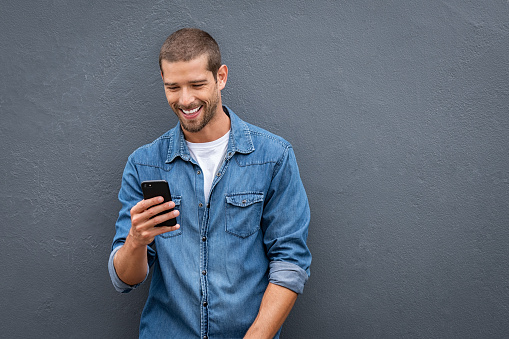 Young man leaning against a grey wall using mobile phone with copy space. Happy casual guy messaging on smartphone on gray background. Cheerful man looking down while typing and reading a message on cell phone.