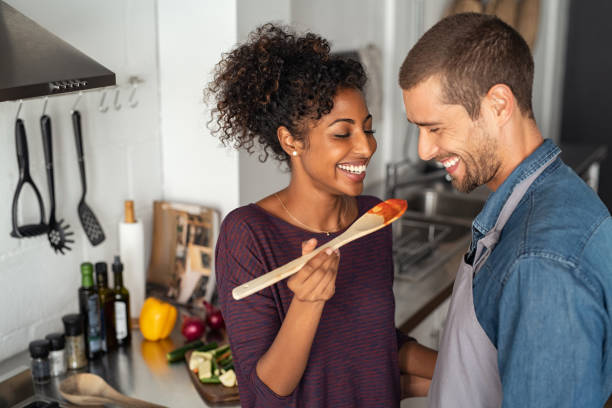 Multiethnic couple tasting food from wooden spoon Young couple tasting tomato sauce while cooking in the kitchen. Cheerful man and smiling woman holding spatula in hand ready to taste red sauce. Multiethnic couple cooking together at home. people preparing food stock pictures, royalty-free photos & images