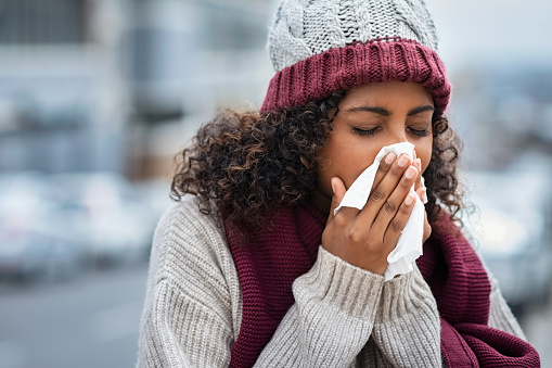 Pretty black woman blowing her nose with a tissue outdoor in winter. Young african woman getting sick with flu in a winter day. Woman with a cold rubs nose with handkerchief.