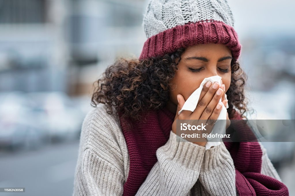 Black woman with cold sneezing outdoor Pretty black woman blowing her nose with a tissue outdoor in winter. Young african woman getting sick with flu in a winter day. Woman with a cold rubs nose with handkerchief. Winter Stock Photo