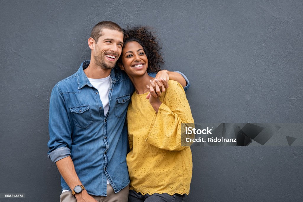 Multiethnic couple in love standing and holding hands Young multiethnic couple in love isolated on grey background looking up and thinking about their future together. Smiling man and african woman in casual hugging and looking away while planning the future. Cheerful couple holding hands while leaning on wall. Couple - Relationship Stock Photo