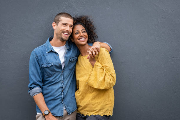 Multiethnic couple in love standing and holding hands Young multiethnic couple in love isolated on grey background looking up and thinking about their future together. Smiling man and african woman in casual hugging and looking away while planning the future. Cheerful couple holding hands while leaning on wall. young couple stock pictures, royalty-free photos & images