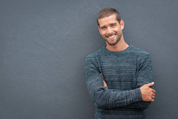 Smiling young man leaning against grey wall Portrait of happy young man leaning against wall isolated on grey background with a big smile. Handsome cheerful guy in winter clothes on gray wall looking at camera. Stylish man wearing sweater with crossed arms standing against wall with copy space. cardigan sweater stock pictures, royalty-free photos & images