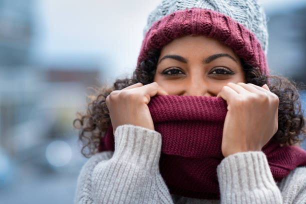 Beautiful woman hiding face in woolen scarf Closeup face of happy african girl holding woolen scarf with hands over nose to protect from the frost. Portrait of beautiful young black woman in warm winter knitted clothes covering her face and looking at camera. warm clothing stock pictures, royalty-free photos & images