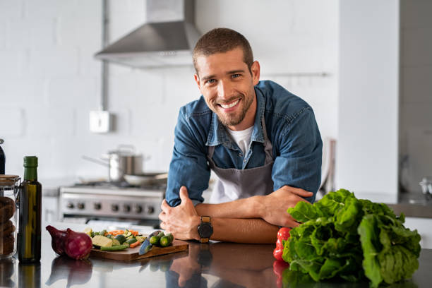 Happy man ready to cook in kitchen Handsome young man leaning on kitchen counter with vegetables and looking at camera. Smiling man wearing apron while preparing food at home. Portrait of happy casual guy leaning on steel counter in the kitchen with vegetables and ingredients on it. home cooking stock pictures, royalty-free photos & images