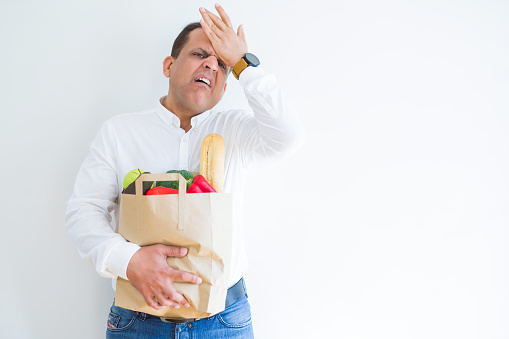 Middle age man holding groceries shopping bag over white background stressed with hand on head, shocked with shame and surprise face, angry and frustrated. Fear and upset for mistake.