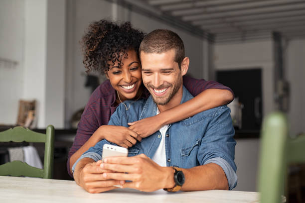 Happy couple looking at phone together Smiling young couple embracing while looking at smartphone. Multiethnic couple sharing social media on smart phones while sitting at table. Smiling african girl embracing from behind her happy boyfriend while using cellphone and laughing. black man white woman couple stock pictures, royalty-free photos & images