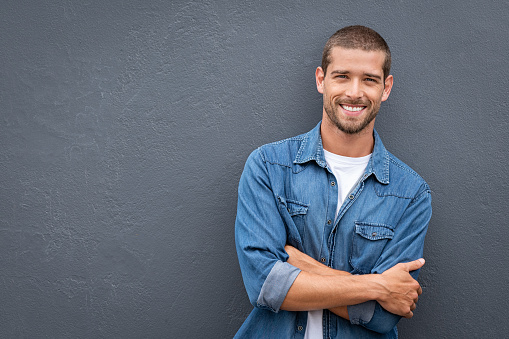 Portrait of handsome young man in casual denim shirt keeping arms crossed and smiling while standing against grey background. Stylish and confident guy leaning against gray wall with copy space. Cheerful friendly man laughing and looking at camera.