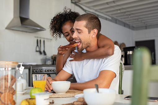 African american woman embracing from behind her boyfriend and feeding him with brown bread and jam. Multiethnic couple enjoying breakfast at home. Beautiful laughing woman feeding young man in kitchen and having fun together in the morning.