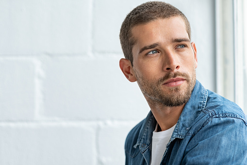 Thoughtful young man sitting near window and looking away. Handsome pensive guy lost in deep thoughts while looking outside the window. Closeup face of man with beard and casual cloth contemplating, copy space.