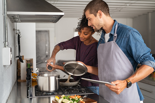 Young man in apron helping african woman to prepare lunch. Happy girl adding salt in pot for pasta while guy hold the lid up. Multiethnic couple cooking together with the help of a digital tablet for the recipe.