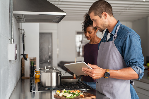 Man and beautiful woman preparing lunch while looking recipe from digital tablet. Young multiethnic couple using computer while cooking in kitchen at home. Husband and wife cooking pasta together according to a recipe on a laptop.