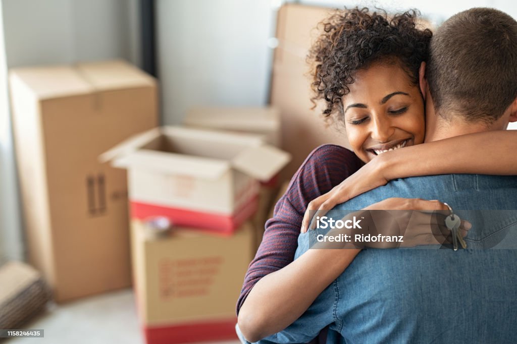 Woman hugging man and holding home keys Young african woman holding home keys while hugging boyfriend in their new apartment after buying real estate. Lovely girl holding keys from new home and embracing man. Happy couple in their apartment around cardboard boxes. Moving House Stock Photo