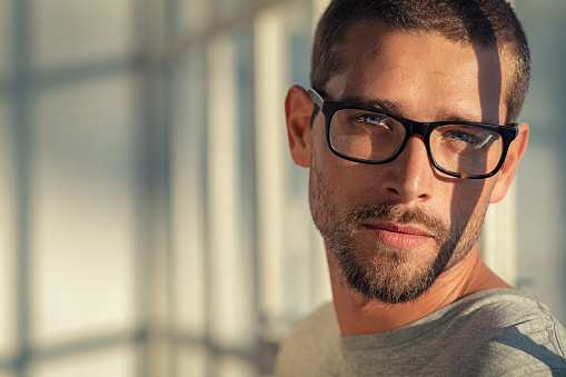 Portrait of stylish young man wearing eyeglasses at home. Closeup face of handsome guy wearing spectacles with sunlight and shadow reflection on face. Portrait of satisfied cool man in casual looking at camera with copy space at sunset.