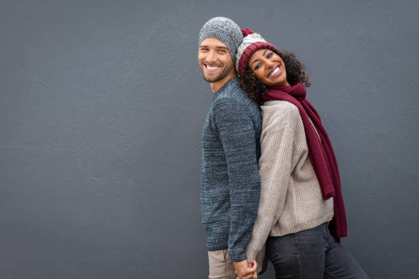 Happy couple in winter clothes leaning against back Young multiethnic couple holding hands while standing and leaning on each other back. Happy man and black woman standing back to back and smiling against grey wall with copy space. Cheerful guy and beautiful woman in warm clothes looking at camera isolated. winter fashion stock pictures, royalty-free photos & images