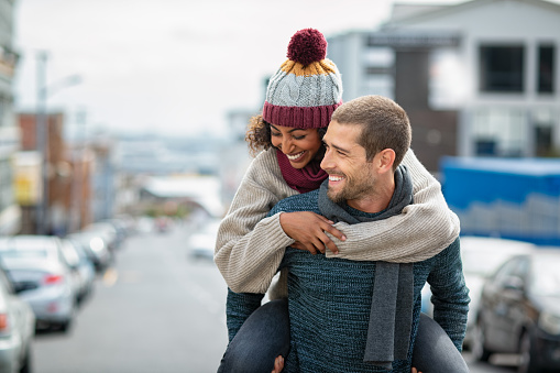 Smiling man giving piggyback ride to woman in the city. Young multiethnic couple in cold clothes walking in street and having fun. Cheerful girlfriend with wool cap and boyfriend in sweater enjoying winter together outdoor.
