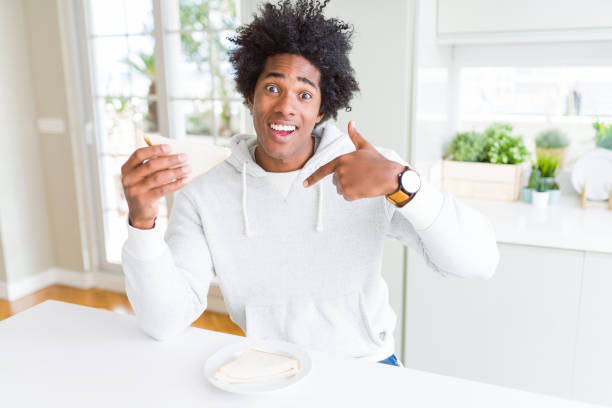 homme d'afro-américain mangeant le sandwich fait à la main à la maison avec le visage de surprise pointant le doigt à lui-même - bon appetite photos et images de collection
