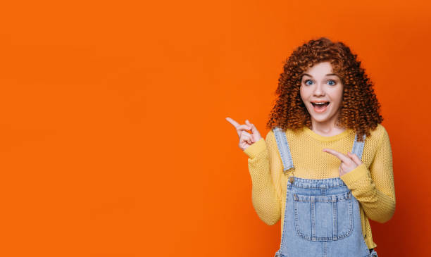 istock curly red-haired woman making surprised face and pointing hands into empty orange background. Wow it amazing 1158247300