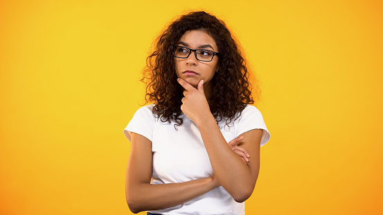 Young biracial woman in eyeglasses thinking about decision, yellow background