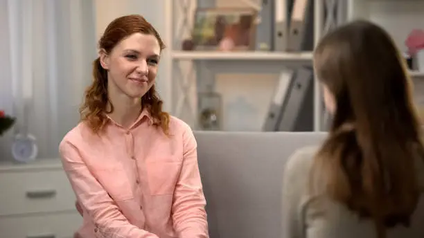 Pretty young woman smiling to female friend, talking on sofa, companionship