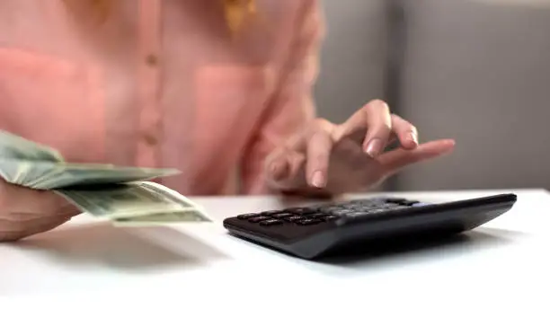 Young lady counting expenses using calculator, holding dollars in hand, income