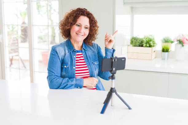istock Senior woman doing video chat using smartphone camera with a big smile on face, pointing with hand and finger to the side looking at the camera. 1158253294