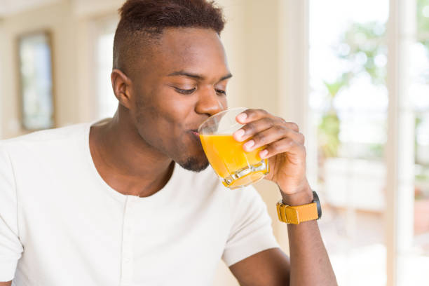 Handsome young african man drinking a glass of fresh natural orange juice enjoying fruit refreshment Handsome young african man drinking a glass of fresh natural orange juice enjoying fruit refreshment juice drink stock pictures, royalty-free photos & images