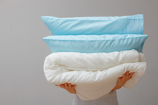 Woman holding a quilt and pillows on a background of gray wall. Stack bedding for sleep. Household.