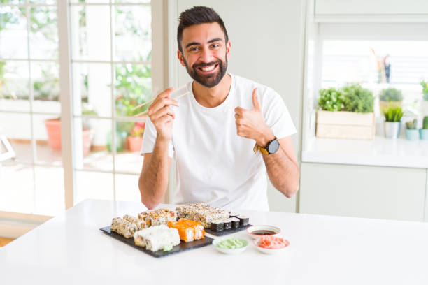 istock Handsome hispanic man eating asian sushi using chopsticks happy with big smile doing ok sign, thumb up with fingers, excellent sign 1158260057