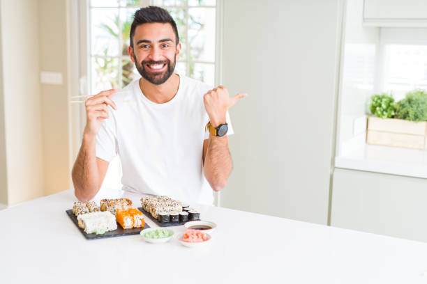 istock Handsome hispanic man eating asian sushi using chopsticks pointing and showing with thumb up to the side with happy face smiling 1158260063