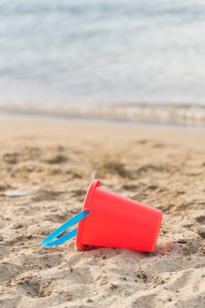 istock Child's red sand pail lying on sandy beach with water in background 1158267988