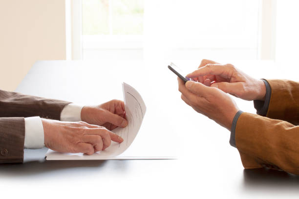 istock Hands from two businessmen in conversation by a desk. One pointing in some papers and the other texting on a cell phone. Negotiating business or a job interview. - Image 1158292964