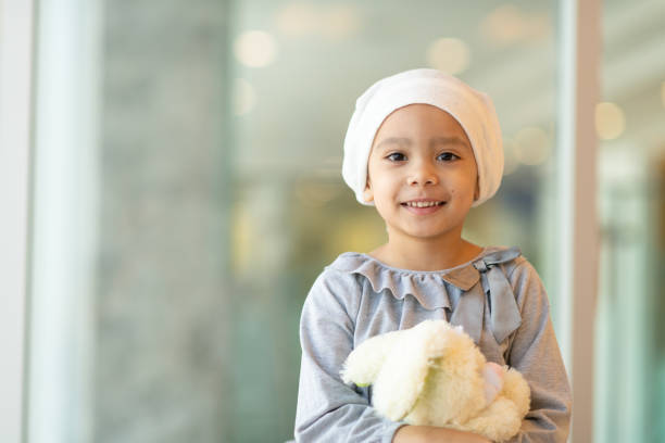 Portrait of a young ethnic girl with cancer A beautiful little girl with cancer takes a break from treatment. She is standing near a large bay of windows in the hospital's corridor. The girl is wearing a headscarf and is holding a stuffed rabbit toy. She is looking at the camera with a big smile. cancer illness stock pictures, royalty-free photos & images