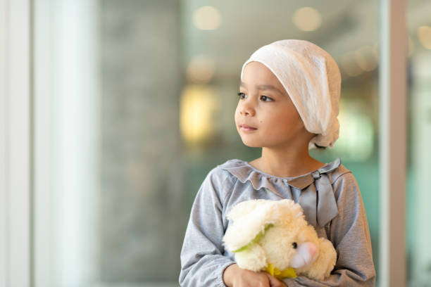 Portrait of a young ethnic girl with cancer A beautiful little girl with cancer takes a break from treatment. She is standing near a large bay of windows in the hospital's corridor. The girl is wearing a headscarf and is holding a stuffed rabbit toy. She is looking out the window with a peaceful expression. cancer cell stock pictures, royalty-free photos & images