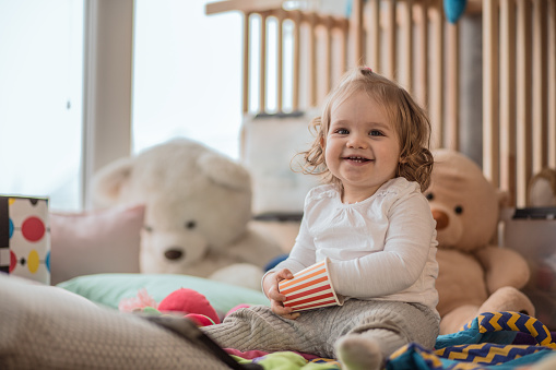 Copy space shot of smiling baby girl playing with a disposable cup while sitting among her toys on the floor of her playroom.