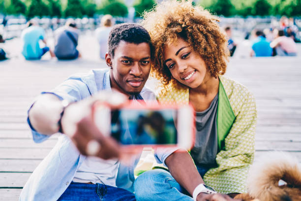 istock Selective focus on young afro american friends posing while talking picture on modern smartphone camera sitting with dog in park, dark-skinned couple using mobile for making photo resting together 1158305049
