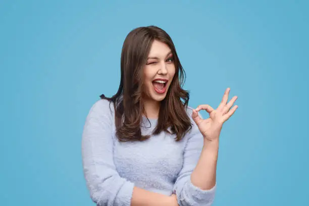 Delighted female in soft sweater showing OK gesture and winking while standing against blue background and looking at camera