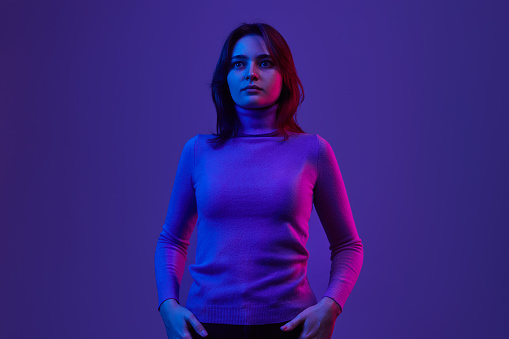 Pretty young female keeping hands in pockets and looking away while standing under bright fluorescent light against violet background