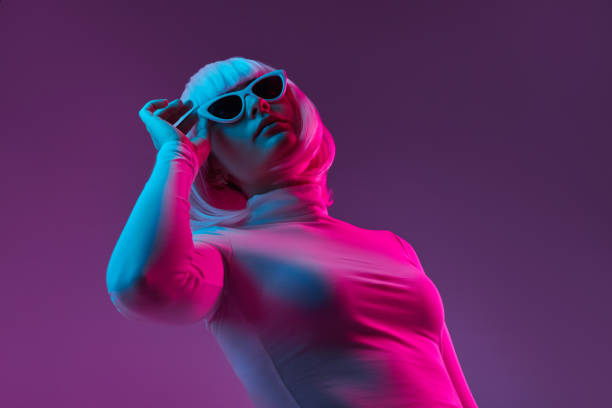 Trendy female in sunglasses looking up Cyberpunk young woman in wig and turtleneck sweater adjusting stylish shades and looking up while standing under neon light against violet background turtleneck photos stock pictures, royalty-free photos & images