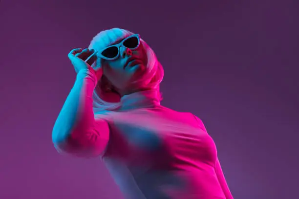 Cyberpunk young woman in wig and turtleneck sweater adjusting stylish shades and looking up while standing under neon light against violet background